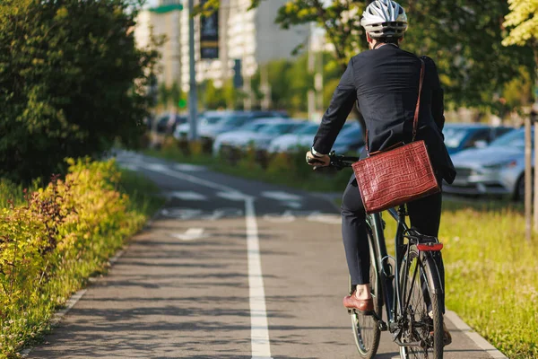 Elegant entrepreneur in smart casual cycling on bicycle track in warm sunny day. Back view of tall male manager with brown leather laptop bag riding bike in sleeping area of city. Concept of activity.