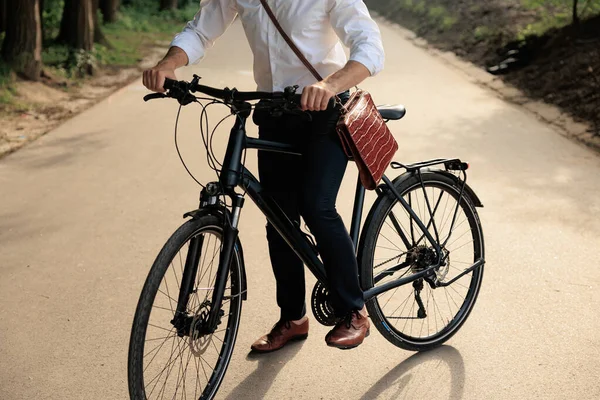 Stylish man with leather briefcase, commuting to work in summer morning. Crop view of unrecognizable male businessman in white shirt starting riding on bike to office outdoors. Concept of lifestyle.