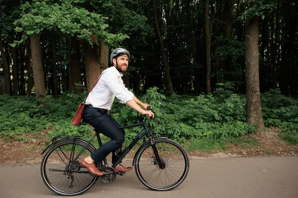 Smiling dark haired businessman on bike, riding along wooded park outdoors. Side view of man wearing smart casual with helmet, enjoying nature, while cycling to work at daytime. Eco-friendly concept.