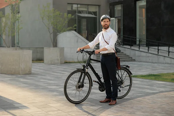 Young businessman commute with bicycle walking home from work in city. Side view of caucasian bearded manager in shirt and helmet standing with modern bike outdoors at daytime. Concept of lifestyle.