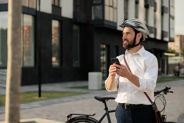 Handsome young man with bike and mobile phone standing on street after workday. Portrait of confident bearded businessman in helmet using cell phone, while looking away in city. Concept of lifestyle.