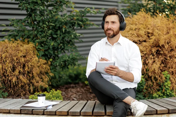 Handsome freelancer in headphones sitting on bench outdoors and writing business plan in notebook. Caucasian male thoughtfully looking aside while working in quiet place.