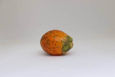 Yakından bakıldığında olgun bir betel fındığı, belirgin turuncu rengi, desenli cildi ve temiz, açık arkaplana karşı doğal yeşil gövdesiyle sergileniyor..