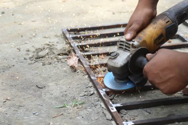 Koyu tenli bir zanaatkar açık havada elektrikli aletle kahverengi metal çerçeveyi düzleştiriyor çimento yüzeyinde altın kıvılcımlar üretiyor..