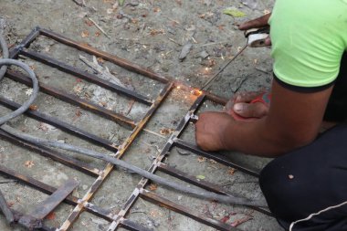 Paslı demir parmaklıkları kaynak yapan Asyalı bir adam, kıvılcımlar uçuşuyor, koruyucu el giysisi ve yeşil şort giyiyor. Metalin etrafında bitki örtüsü olan görülebilir bir yer..