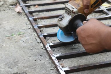 Bir işçi, el kameralı bir değirmen kullanarak kaynaklı metal çerçeveyi düzleştiriyor. Metal işçiliği sırasında kıvılcımlar görünür. Araç ve ellere yakın çekim.