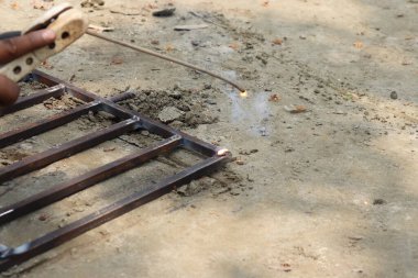 Bir işçi, çubuklar kullanarak paslı, kare bir metal çerçeve kaynak yapar; kıvılcımlar koyu tenli bir el tarafından tutulan eklem yayılır cesur bir yüzey üzerinde bir klemp kullanarak.