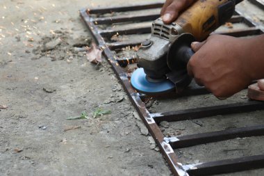Metal çerçeve öğüten Asyalı bir adamın açık havada parlak kıvılcımlar saçan görüntüsü. Demircilik, arka bahçedeki çalışma alanında beceri ve hassasiyet gösteriyor..