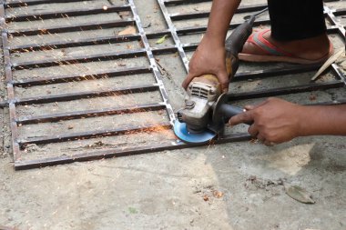 Asyalı bir adamın el aletiyle metali öğüttüğü yakın çekim; iş sırasında kıvılcımlar uçuşuyor, dışarıda yere seriliyor.