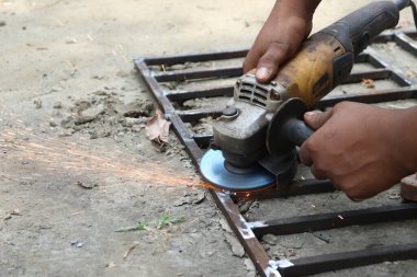 Yakından çekilen görüntülerde, koyu tenli bir insan metal çerçevede öğütücü kullanırken kıvılcımlar uçuşuyor ve öğütme işlemine odaklanıyor..
