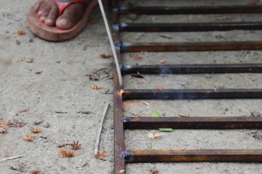 Metal çerçeve kaynaklanırken kıvılcımlar ve duman ortaya çıkar. Koyu tenli bir insan kısmen görülebilir, ayağı sandalette. Toprakta organik madde var..