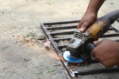 Çelik çerçeve üzerinde bir açı öğütücü kullanan koyu tenli metal işçisinin yakın çekimi, açık hava ortamında kıvılcımlar üretiyor. El ve araç ayrıntılarını gösterir.