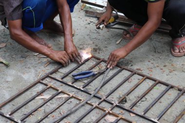 Yakın çekim, iki Asyalı adama odaklanmış yerde kıvılcımlar saçan metal bir şebeke ve çalışma alanının yakınında mavi bir kerpeten..