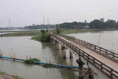 Su bitkisi, boru hattı ve Bangladeş kırsalındaki uzak kuleyle bulanık bir nehri kaplayan yıpranmış beton bir köprüyü gösteren manzara..