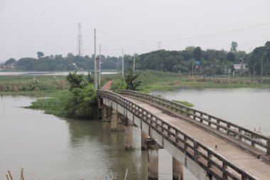 Bereketli ağaçlarla çevrili sakin bir nehrin üzerindeki beton köprü. Bulutlu bir gökyüzünün altında, arka plandaki uzak yapı doğal bir manzara sağlıyor..