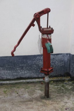 Yıpranmış dokuyu ve kullanışlı kırmızı demir bir su pompasını yakalar, sapını, yeşil borusunu ve sağlam yapısını gösterir. Açık havada, kırsal cazibe uyandırır..