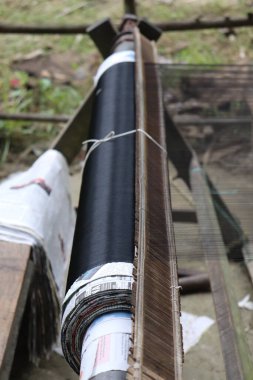 Yan görünüş, dokuma tezgahının geleneksel yapısı üzerine siyah iplik ve gazeteyle dokunmuş dokuyu odaklıyor, işçiliği ve dokuma sürecini vurguluyor.