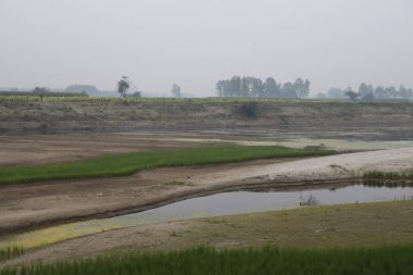 Ufukta bir kişinin yürüdüğü, bulutlu bir gökyüzünün altında nehir, yeşil çimenler ve uzak ağaçların bulunduğu sakin kırsal bir manzara.