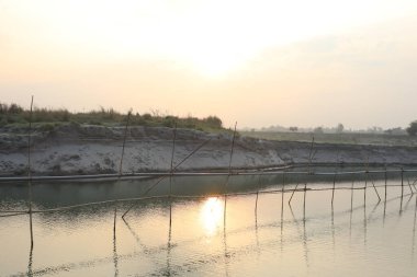 Gün batımını örülmüş bambu destekli geniş bir nehirde yansıtıyor. Altın ışık sakin bir atmosferi resmediyor..