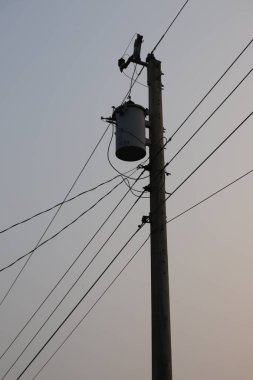 Yukarı doğru ateş eden, koyu renk siluette uzun tahta bir elektrik direği yakalıyor. Gri transformatör, bağlı kablolar ve sessiz arka planda birden fazla siyah elektrik hattı var..