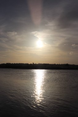 Güneş ışığı, bulutlarla dolu bir gökyüzünün altında uzanan ağaçlardan oluşan ince bir çizgiyle güneşi yansıtan sakin nehir suyunda parıldayan bir yol yaratır..