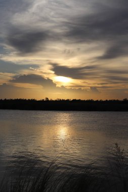 Gün batımında bir nehrin sakin güzelliğini yansıtan sakin bir görüntü. Güneş ışığı, suyun üzerine parlak bir ışık saçıyor. Bulutlar olay yerine derinlik katar, çarpıcı bir manzara yaratır..
