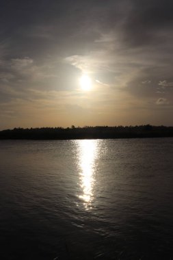 Manzarada, bulutlu bir gökyüzünün altında, sakin bir atmosfer ve doğal bir zarafet sunan ılık güneşin, sakin bir nehre parlak bir şekilde yansıması görülüyor..