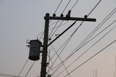 Görüntü, silindirik transformatörlü bir elektrik direğine odaklanıyor. Yumuşak, puslu bir gökyüzüne karşı farklı yönlerde çalışan birkaç elektrik telini vurguluyor..