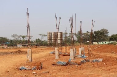 Görüntü, arka planda açık bir gökyüzü ve bitki örtüsü ile kırmızı toprak üzerindeki üst kümeden çıkan paslı metal çubuklu beton destek sütunlarını gösterir.