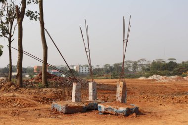 Fotoğrafta, tuğlalar da dahil olmak üzere kırmızı toprağı ve dağınık döküntüleri bulunan bir inşaat sahasının ortasında inşa edilmiş beton destek yapıları yer alıyor. Ufukta ağaçlar ve binalar var..