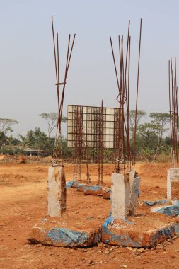 Beton yapılar, paslı metal kirişlerle kırmızımsı toprağın üzerinde duruyor. Bu da arka planda orman olan bir reklam panosunun iskelet yapısına işaret ediyor..