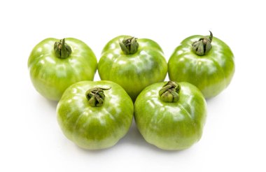 Yakın plan çekimde, pürüzsüz beyaz bir arkaplanda sıralanmış, olgunlaşmamış beş adet yeşil aile yadigarı domates yer alıyor. Şekilleri ve dokuları taze, organik bir izlenim veriyor..