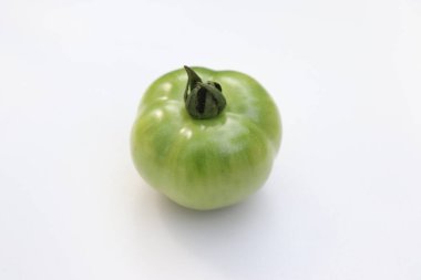 Tek, canlı yeşil bir domates, beyaz bir arka planda duruyor. Görüntü domatesin olgunlaşmamış rengini, pürüzsüz dokusunu ve kök detaylarını vurgular..