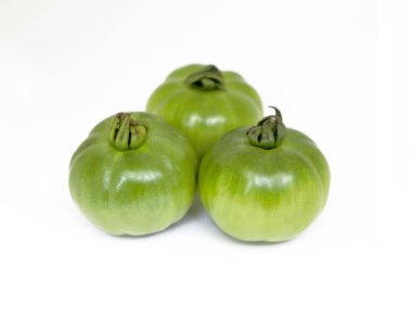 Yakın plan çekimde birbirine yakın konumda duran üç taze, olgunlaşmamış yeşil domates canlı renklerini ve eşsiz şekillerini ön plana çıkaran sert beyaz bir zemin üzerine kurulmuş..