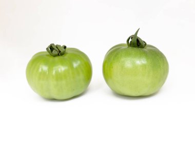 Bu yakın çekim stüdyo çekiminde iki taze, olgunlaşmamış, kökleri olan yeşil domatesler var, dikişsiz beyaz bir zemine yakın konumdalar, ham dokularını geliştiriyorlar..