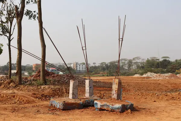 Fotoğrafta, tuğlalar da dahil olmak üzere kırmızı toprağı ve dağınık döküntüleri bulunan bir inşaat sahasının ortasında inşa edilmiş beton destek yapıları yer alıyor. Ufukta ağaçlar ve binalar var..