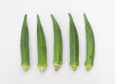 Beş taze, parlak yeşil bamya kabuğu, temiz, beyaz bir yüzey üzerinde sıralanır. Bu, onların dokusunu ve rengini vurgulayan basit ve doğal bir düzenleme sunar..