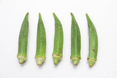 Bu resim, doğal dokularını ve renklerini vurgulayarak el değmemiş beyaz arkaplana karşı bir sırada dizilmiş taze seçilmiş beş yeşil bamya kabuğunu içeriyor..