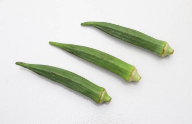 Üç yeşil, taze bamya kabuğu temiz beyaz bir yüzeyde soldan sağa çapraz hizalanır. Sebzeler dokuda ve renkte doğal kusurları gösteren ufak farklılıklar sergiliyor..