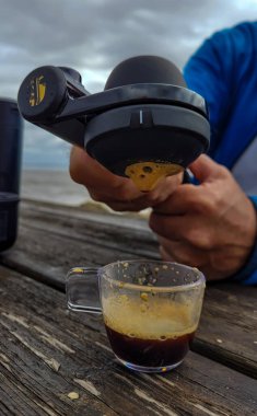 Erkeklerin elleri seyahat kahve makinesini tutuyor. Kahve makinesinin içinden taze ve güçlü bir espresso akıyor. Köpükle birlikte ahşap bir masada bir fincana dökülüyor. 29 Eylül 2022, Talais, Fransa
