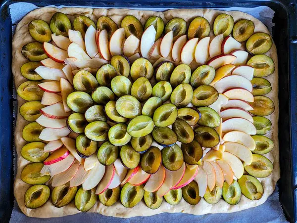 stock image Fresh witched plums halves and slices of apples are located on yeast dough. Fruit cake filling, top view. Preparation of ingredients for baking a cake with fruits.