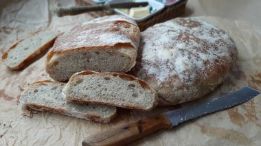 İştah açıcı ev yapımı ciabatta, parça parça kesilmiş, pişirme kağıdının üzerinde bulunur. Yanında tahta saplı bir bıçak var. Arka planda - tereyağı.