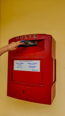 Kadınların eli kırmızı posta kutusuna sarı duvara bir kartpostal atar. Posta kutusunda posta kolleksiyonunun zamanlarıyla ilgili bilgiler var. Klasik İtalyan kırmızı posta kutusu. 20 Eylül 2024. Pomonte, Elba, İtalya