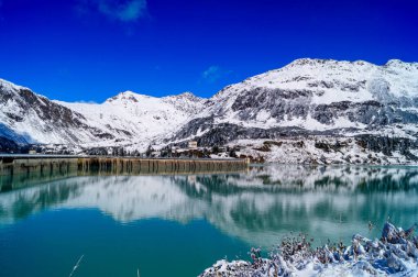 Gök mavisi sularla kaplı, karla kaplı dağlarla çevrili, beton bir baraj ve yansıma manzarası..