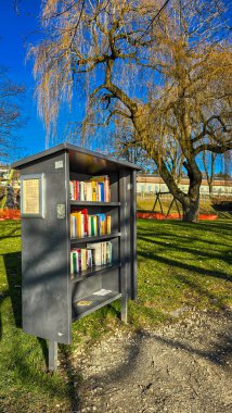 Kitaplar için parkta ücretsiz açık kütüphane. Arka planda çocuklar için yeşil bir çimen, ağaçlar ve oyun alanı var. 26 Aralık 2024. Uberlingen, Almanya.