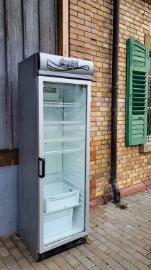 An empty refrigerator with glass doors and a Coca-Cola logo, against the backdrop of a brick and wooden wall. Empty promises. December 12, 2024. Uberlingen, Nussdorf, Germany.