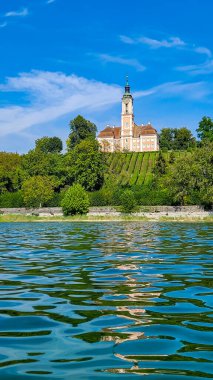 Barok Birnau Bazilikası 'nda Constance Gölü' nün yanından bir görüntü. Ön planda göl, ağaçlar ve üzüm bağları var. Kilise suya yansıtılır. Dikey biçim.