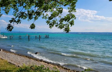 Güneşli bir günde mavi-turkuaz suyu olan güzel bir Constance gölü manzarası. Ön planda, rüzgarın estiği ağaçların dalları. Bodensee, Almanya.