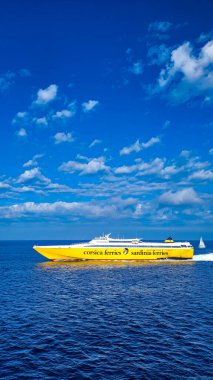Güneşli bir günde Elba Adası 'na giden Korsika Sardunya Feribotları tarafından işletilen feribot. Ön planda koyu mavi bir deniz, arka planda beyaz bir yelkenli. Dikey format. 28 Eylül 2025. Piombino, İtalya.