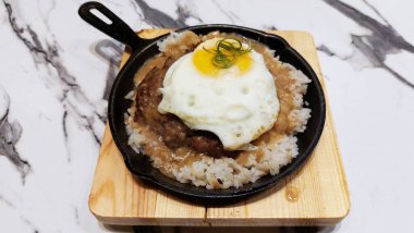 Burger steak patty with white rice and sunny side up egg served in black cooking pan